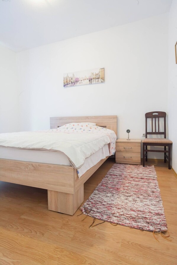 Un dormitorio con cama de madera, una mesita de noche, una silla, una pequeña mesa y un cuadro colgado en la pared.