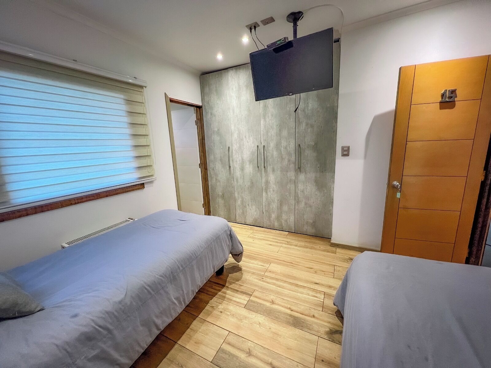 Une chambre avec deux lits simples, une armoire, un téléviseur fixé au mur et une fenêtre avec des stores.