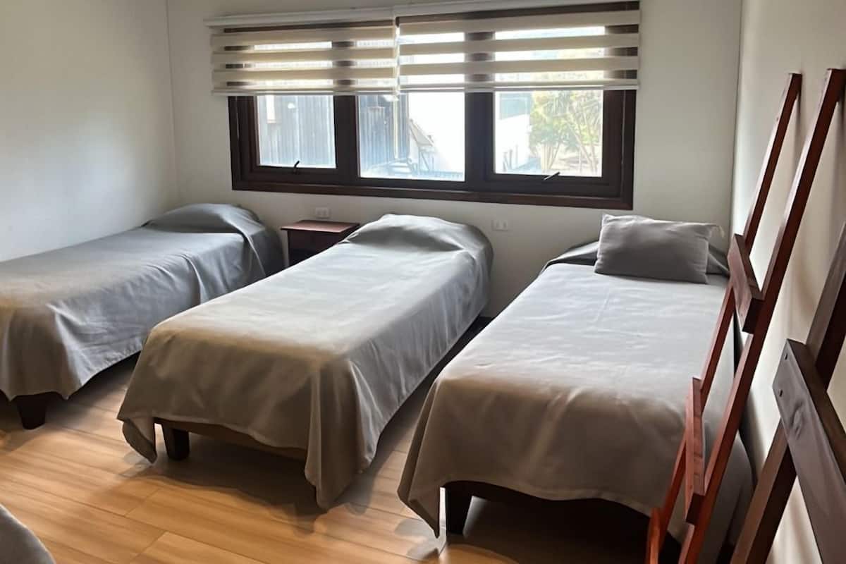 A room with three single beds, wooden flooring, a window with blinds, and a ceiling with recessed lighting.
