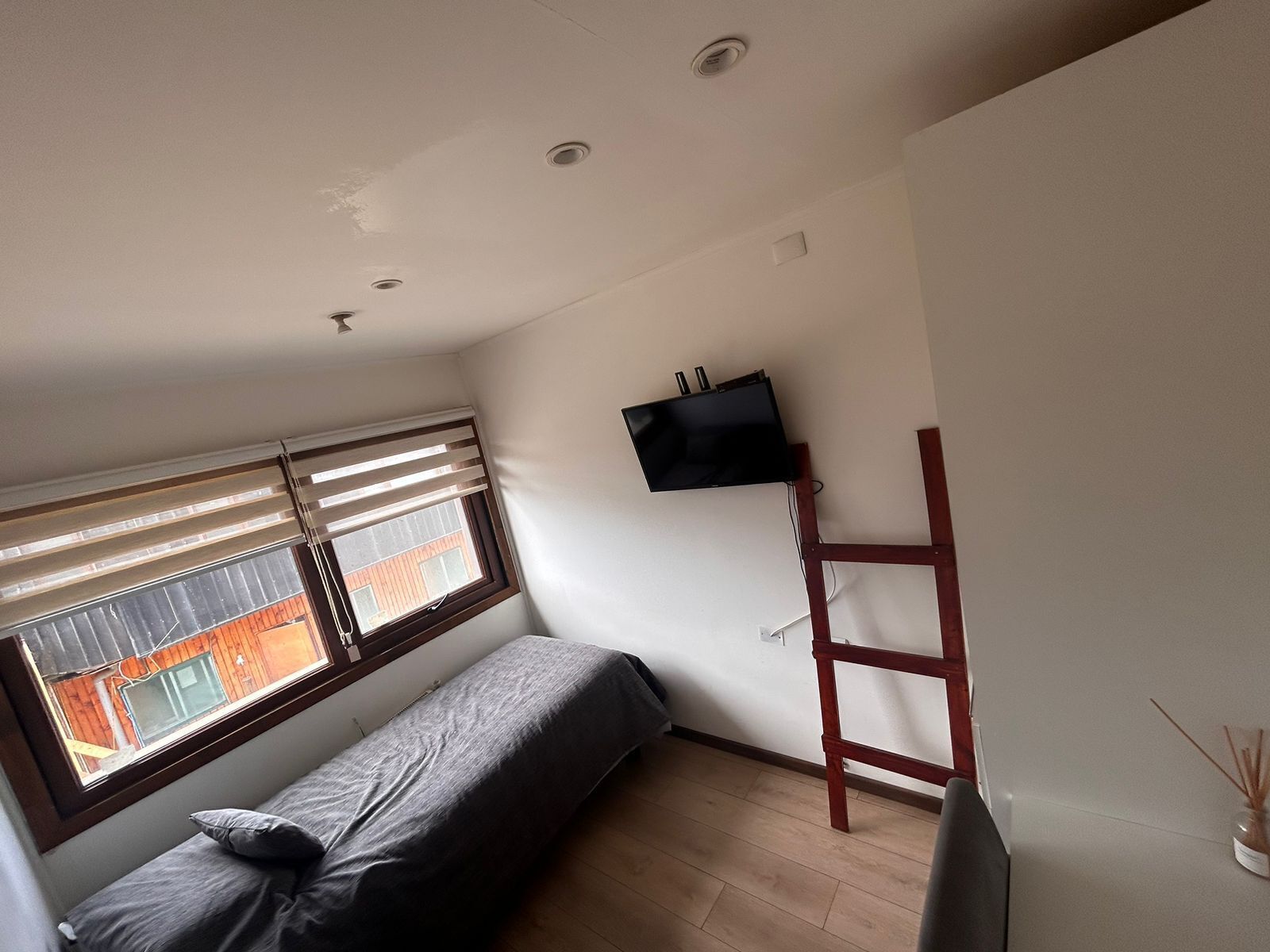 A small, sparsely furnished room with a single bed, a TV mounted on the wall, a window with blinds, and a wooden ladder.
