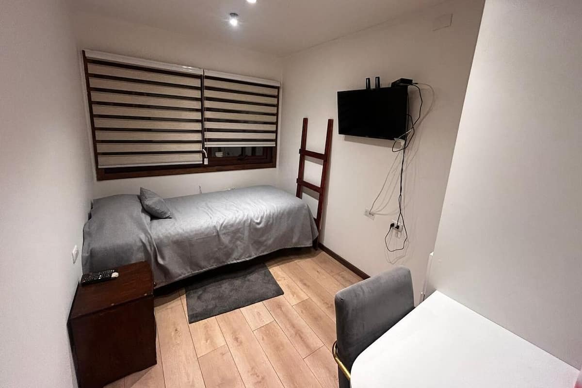 A small, compact bedroom with a single bed, a desk, and a TV mounted on the wall.