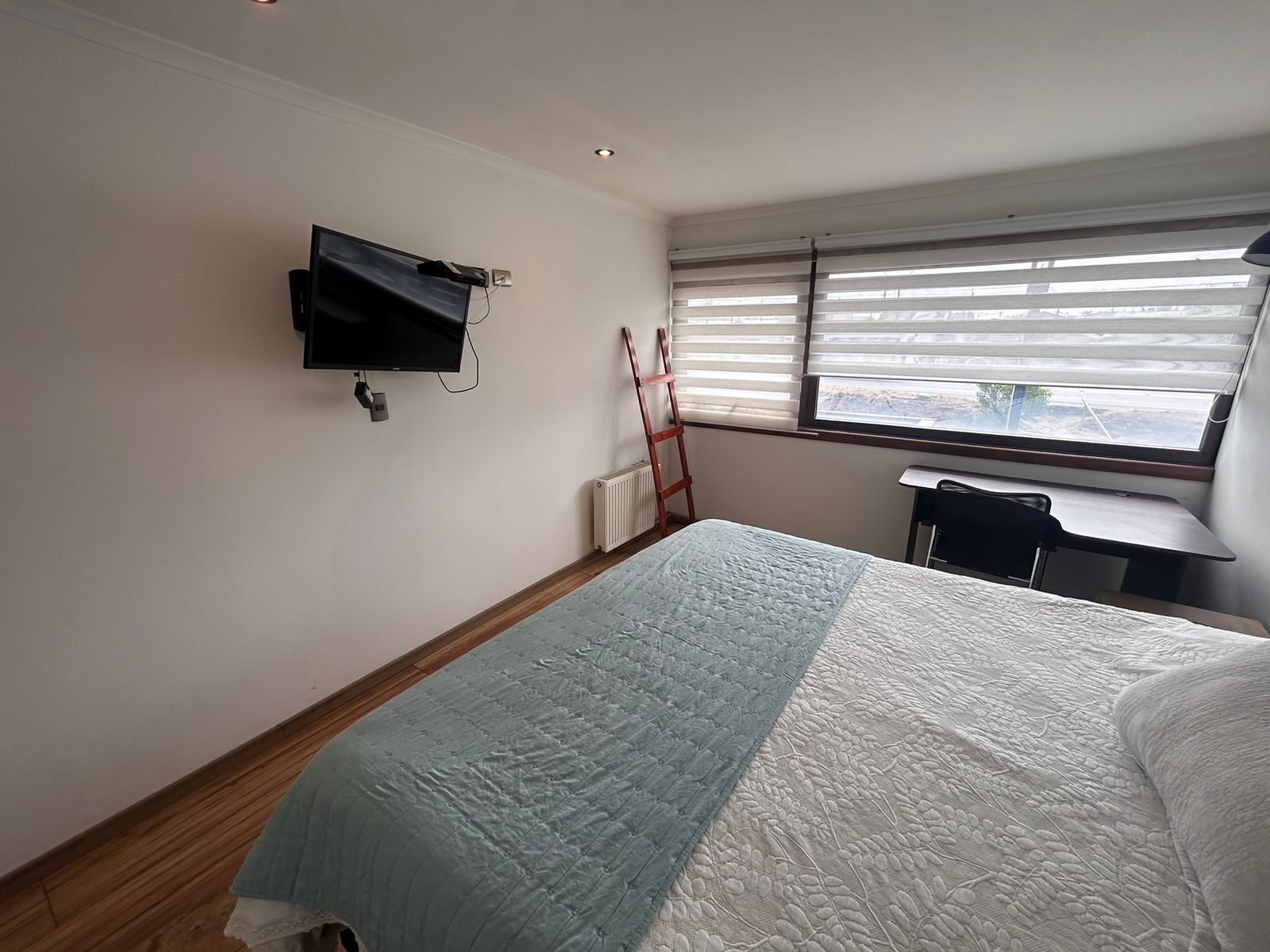 Habitación de hotel con cama, televisor montado en la pared, radiador, ventana con persianas y escritorio con silla.