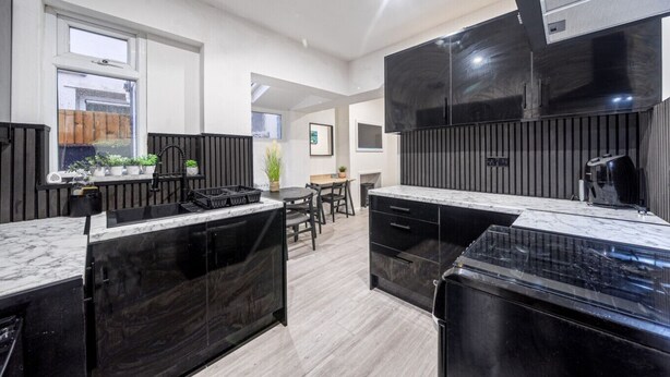 Cozinha moderna com armários pretos, bancada de mármore e fogão embutido. Há uma área de jantar com mesa e cadeiras, e uma janela que proporciona luz natural.