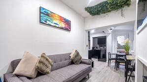 Uma sala de estar moderna com um sofá cinzento, duas almofadas, uma pintura abstrata colorida, uma área de jantar com mesa e cadeiras, e uma cozinha visível através de uma porta aberta.