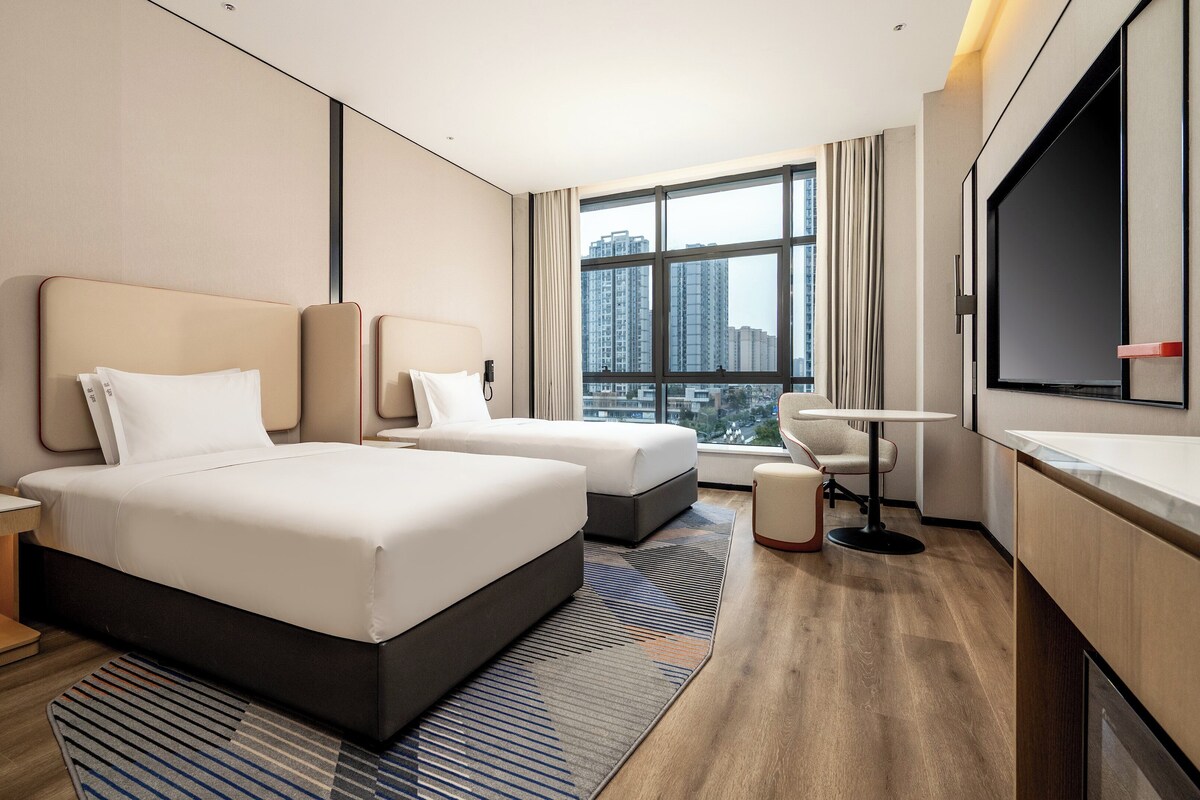 A modern hotel room with two beds, a desk, a chair, a TV, and a large window with a city view.