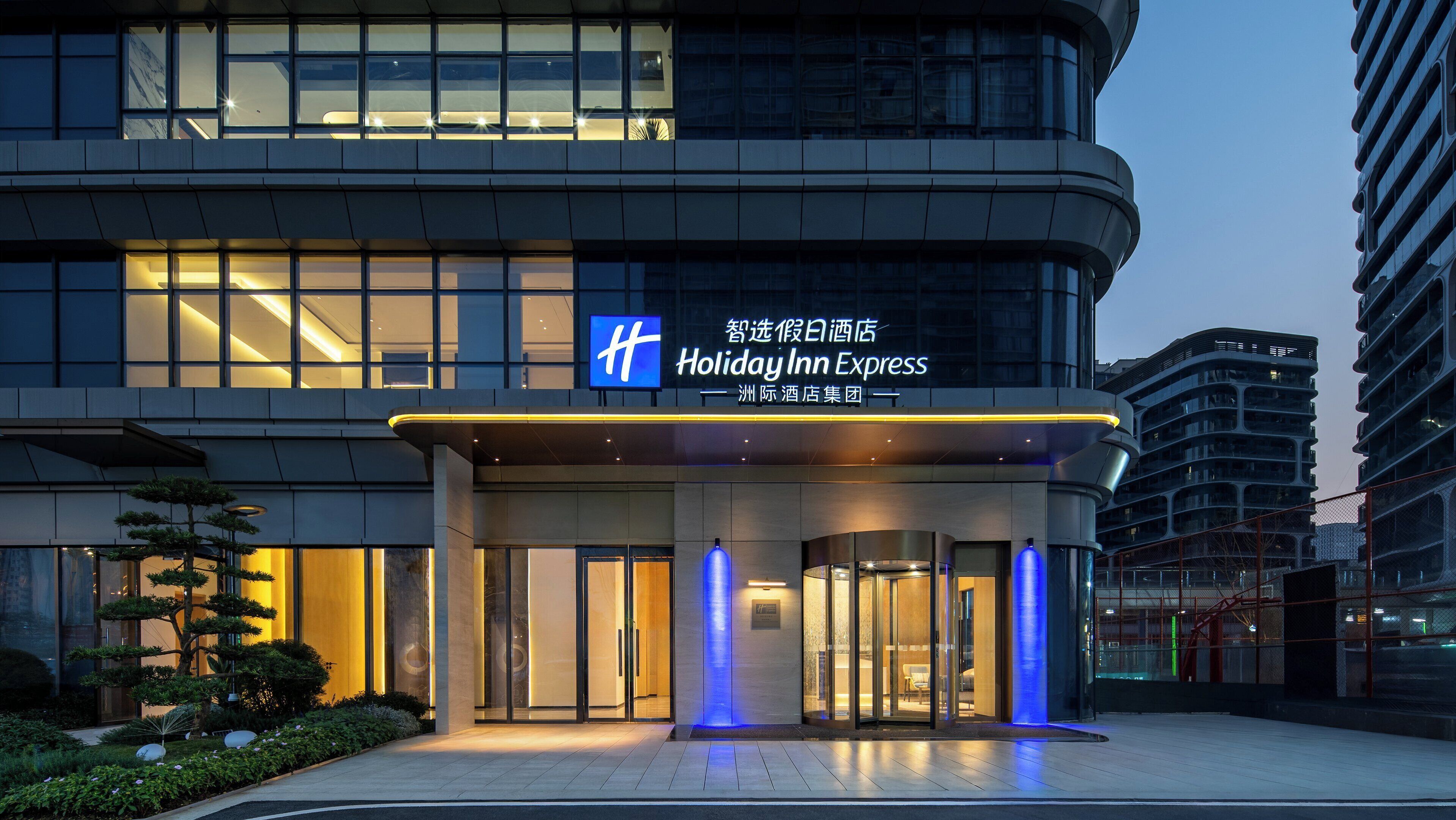 The exterior of the Holiday Inn Express hotel with a modern design, featuring a prominent entrance with glass doors and illuminated blue accents.