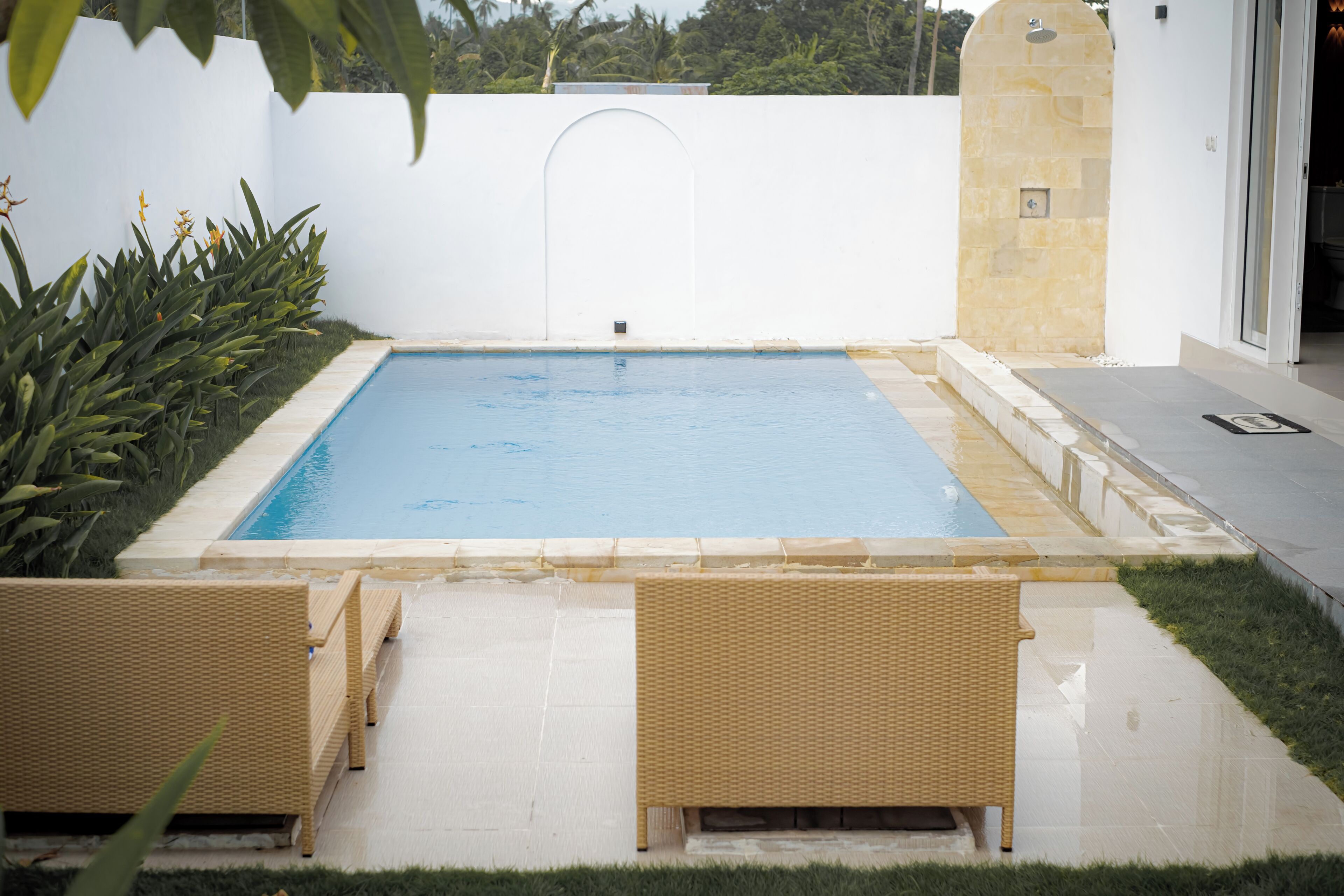 Ein rechteckiger Pool mit weiß gefliestem Rand, umgeben von einer niedrigen Hecke und zwei Korbsesseln.