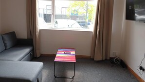 Et værelse med en grå sofa, et farverigt bord, et vindue med gardiner og et fjernsyn på væggen.