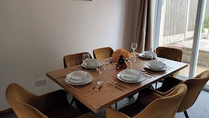 Et spisebord dækket til fire med hvide tallerkner, glas og bestik. Der er en vase med blomster på bordet, og rummet har et stort vindue med gardiner.