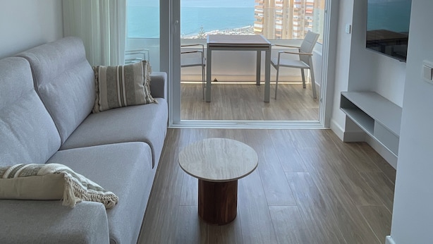 Ein modernes Wohnzimmer mit einer Couch, einem runden Couchtisch und einem Balkon mit Blick auf das Meer und Gebäude.