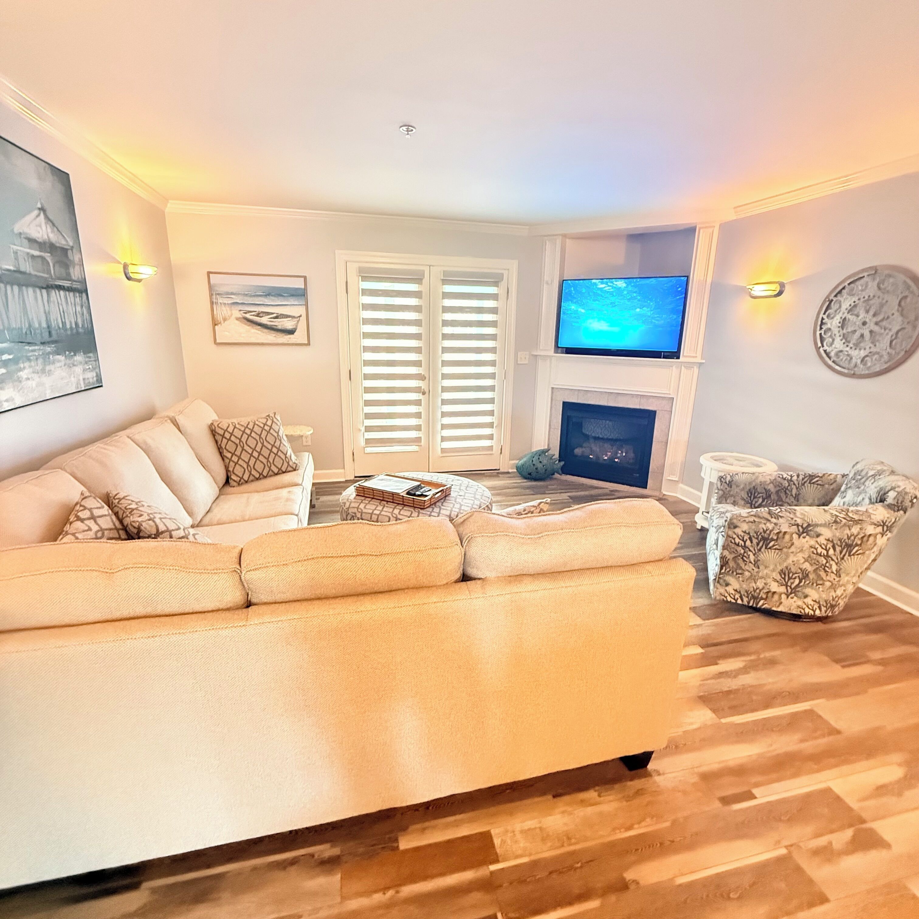 A living room with a large beige sofa, a patterned armchair, a flat-screen TV mounted above a fireplace, and a wall-mounted TV.
