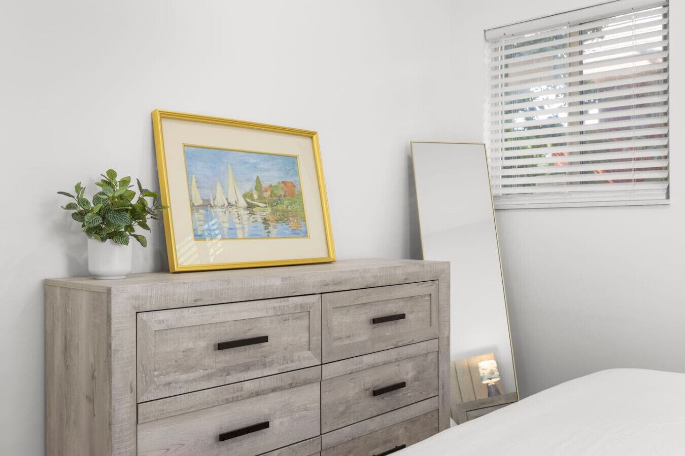 Une chambre à coucher avec un lit, une commode à tiroirs, un tableau encadré, une plante en pot et un miroir.