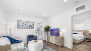 Una habitación de hotel moderna con sofá, un sillón azul, un reposapiés blanco, un televisor sobre un mueble bajo y una cama con una mesita de noche.