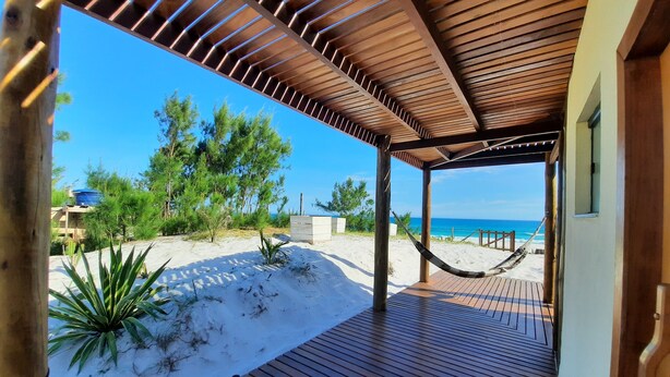 En træterrasse med en hængekøje, der kigger ud over en strand med sand og sparsom vegetation.