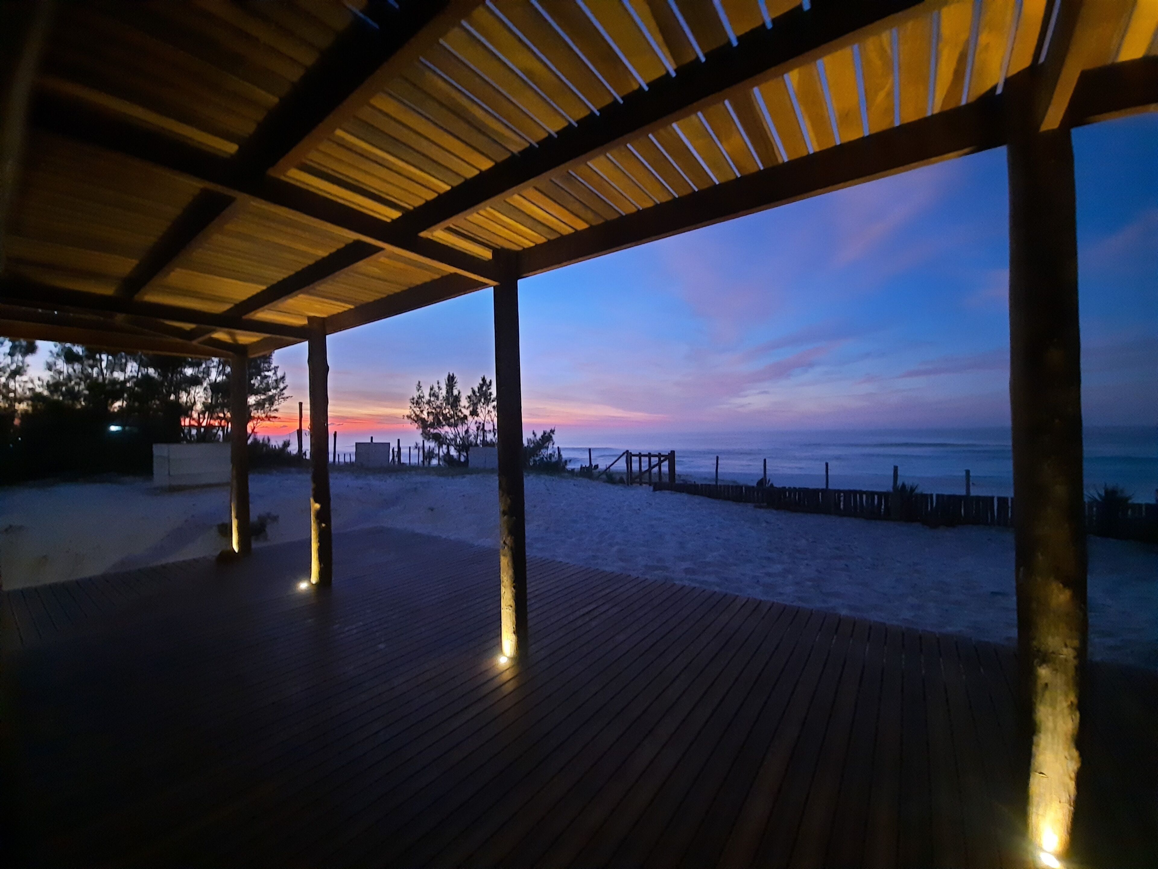 Eine hölzerne Terrasse mit Säulen, die auf einen Strand im Sonnenuntergang blickt.