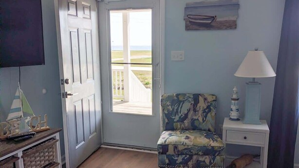 Una habitación con piso de madera, una silla, un televisor, una puerta que da al exterior, una lámpara y una pequeña mesa.