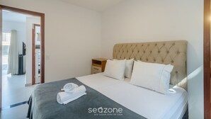 A neatly made bed with a grey bedspread and white pillows, a wooden nightstand with a lamp, and a view of a balcony through an open door. - House in Condo w/ Pool and Barbecue Grill Bme0001 (Porto Seguro)