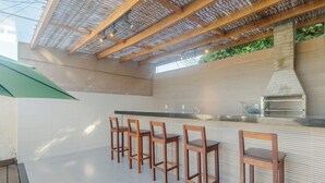 An outdoor kitchen area with a brick wall, a grill, and wooden bar stools. - House in Condo w/ Pool and Barbecue Grill Bme0001 (Porto Seguro)