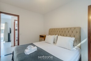 A neatly made bed with a grey bedspread and white pillows, a wooden nightstand with a lamp, and a view of a balcony through an open door. - House in Condo w/ Pool and Barbecue Grill Bme0001 (Porto Seguro)