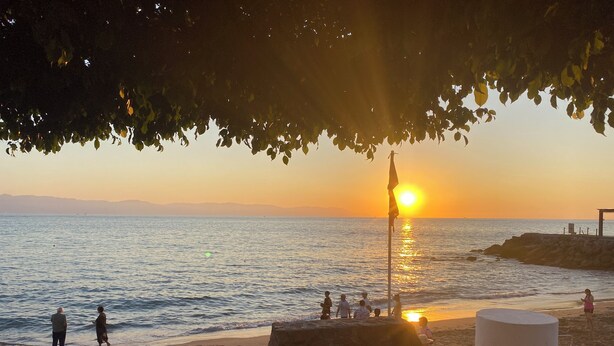 Ein Strand mit Liegestühlen und einem Tisch unter einem Baum, mit Blick auf das Meer und einen Sonnenuntergang.