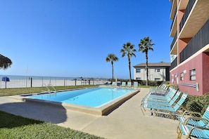 A swimming pool with lounge chairs, palm trees, and a building with balconies. - Emerald Isle 304 - Premier (North Redington Beach)