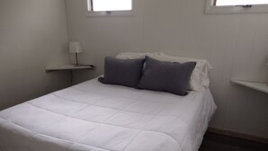 Un dormitorio con una cama blanca, dos almohadas grises, una mesita de noche con una lámpara y una ventana sobre la cama.