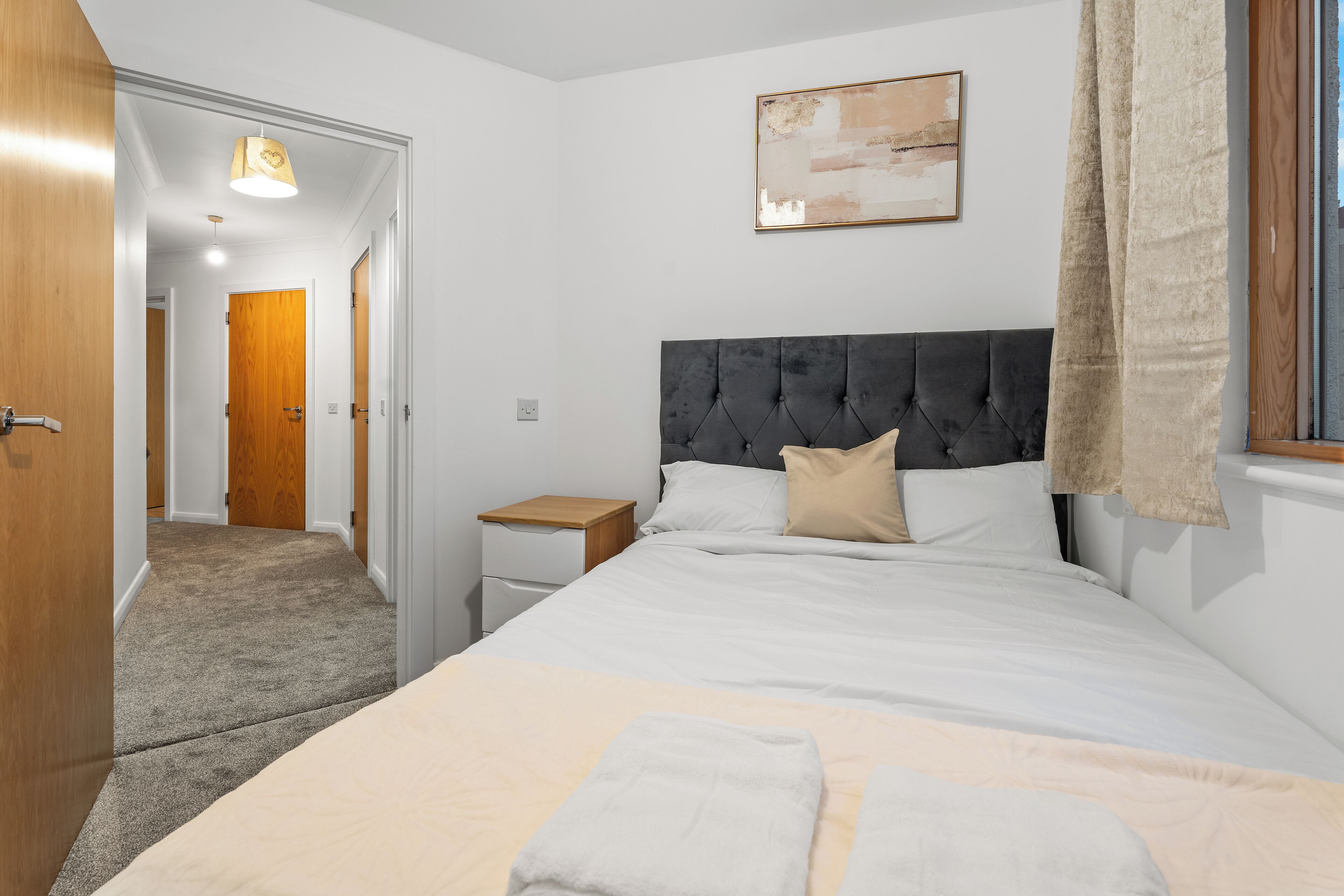 Un dormitorio con una cama grande, una mesita de noche, suelo de madera y un pasillo que conduce a otra habitación.