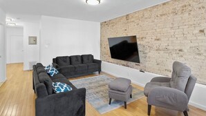 Ein modernes Wohnzimmer mit einer Eckcouch, einem grauen Sessel und einem Flachbildfernseher, der an einer Ziegelwand angebracht ist.