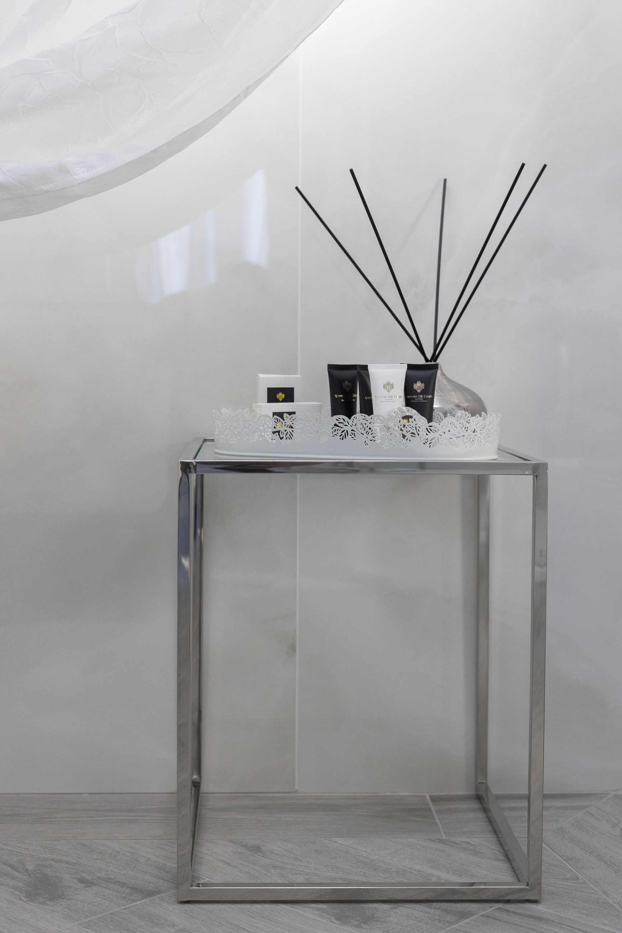 Uma mesa de vidro transparente com base em renda branca, uma pequena caixa branca e dois frascos de perfume pretos.