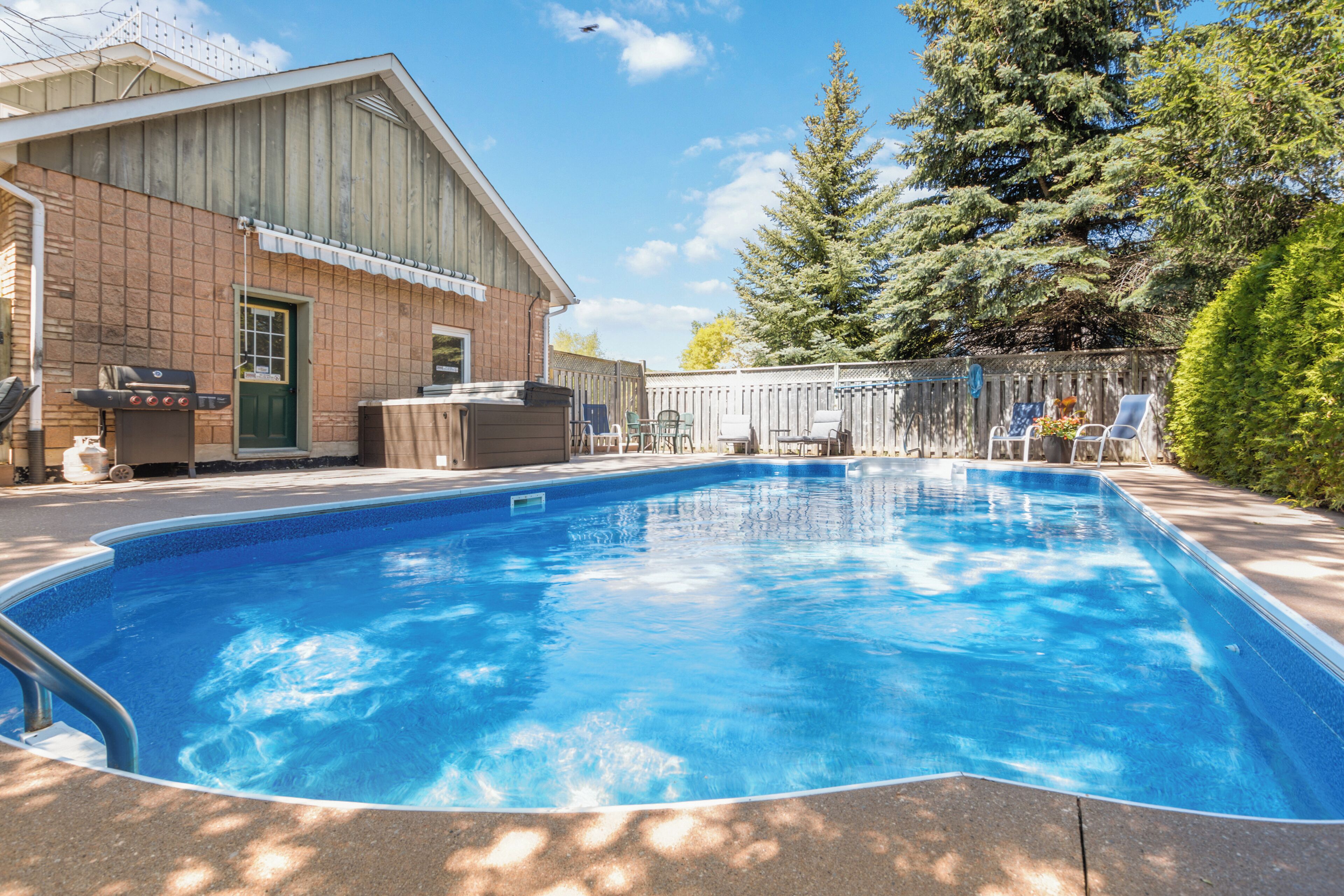 A swimming pool with a wooden deck, a barbecue grill, and a fenced area with chairs.