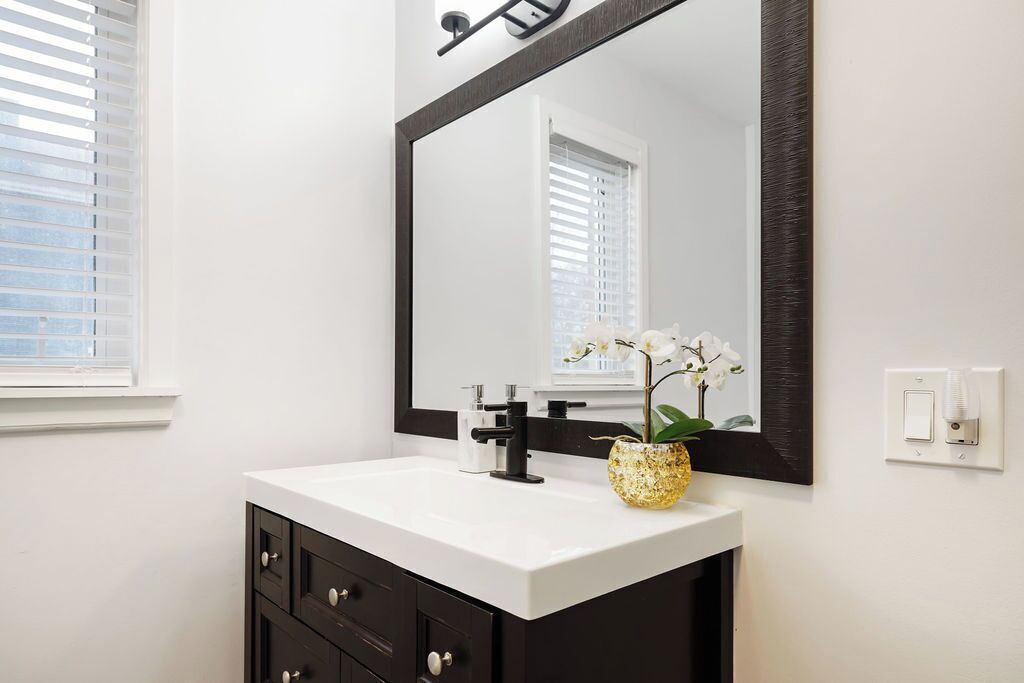 A bathroom with a white vanity, dark cabinets, a large mirror, and a small vase with flowers.