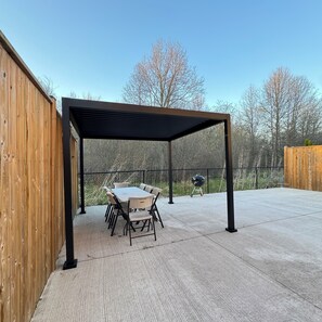 A covered patio area with a table and chairs, a grill, and a wooden fence.
