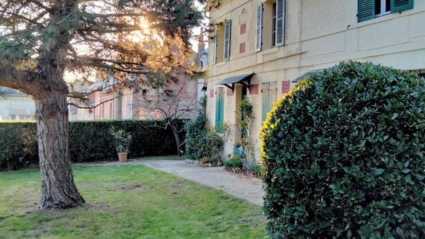 Un bâtiment à deux étages avec un jardin devant, un grand arbre et une haie.