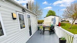 Una terraza techada con una mesa de picnic y sillas, una valla blanca, y una hilera de casas móviles al fondo.