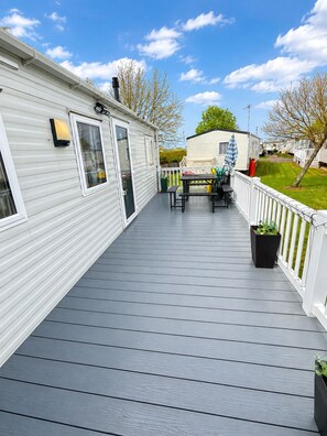 A covered deck with a picnic table and chairs, a white fence, and a row of mobile homes in the background.
