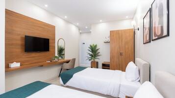 A modern hotel room with two beds, a TV mounted on a wooden panel, a desk with a chair, a wardrobe, and a potted plant.