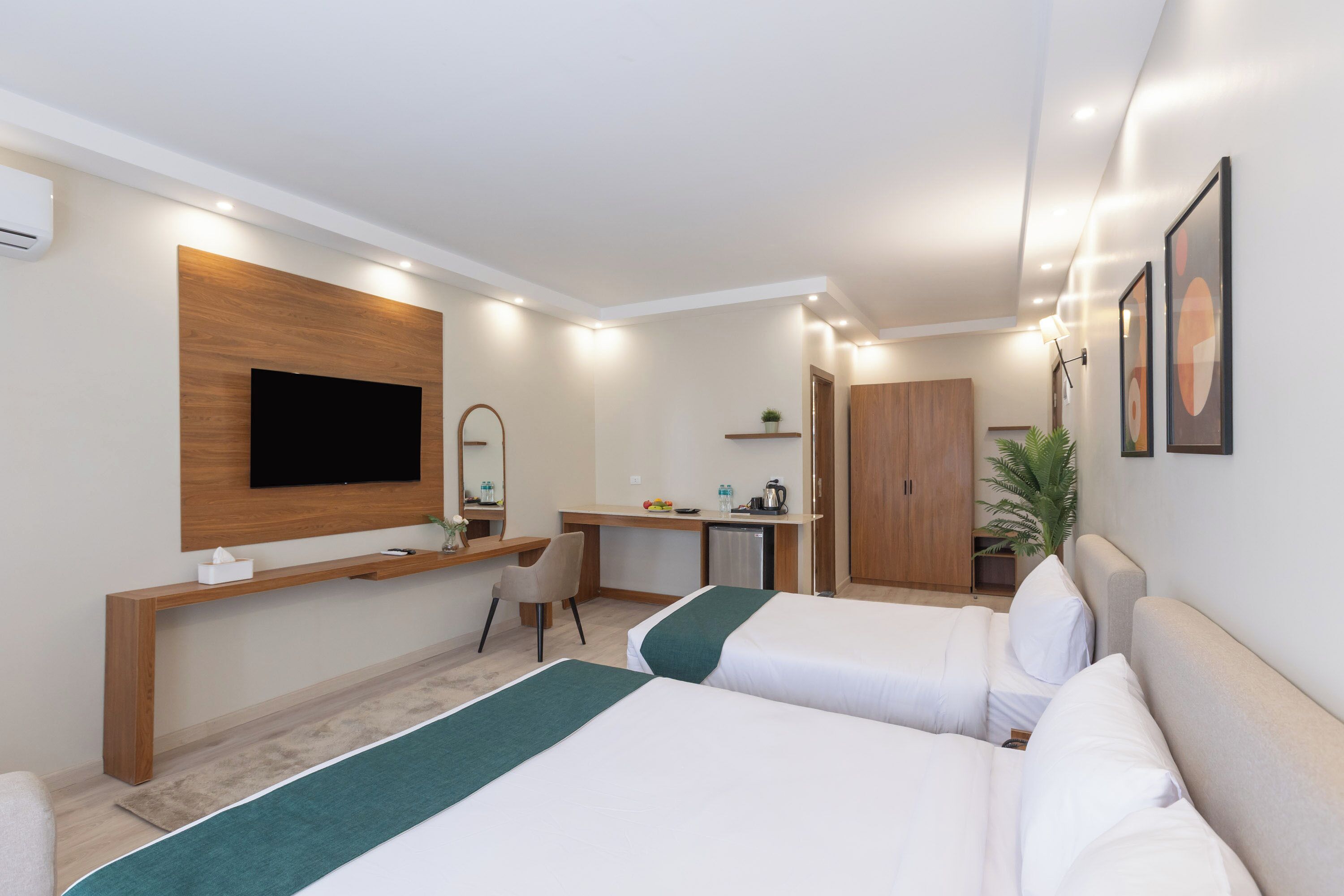 A hotel room with two beds, a desk, a chair, a TV, a wardrobe, and a small plant.