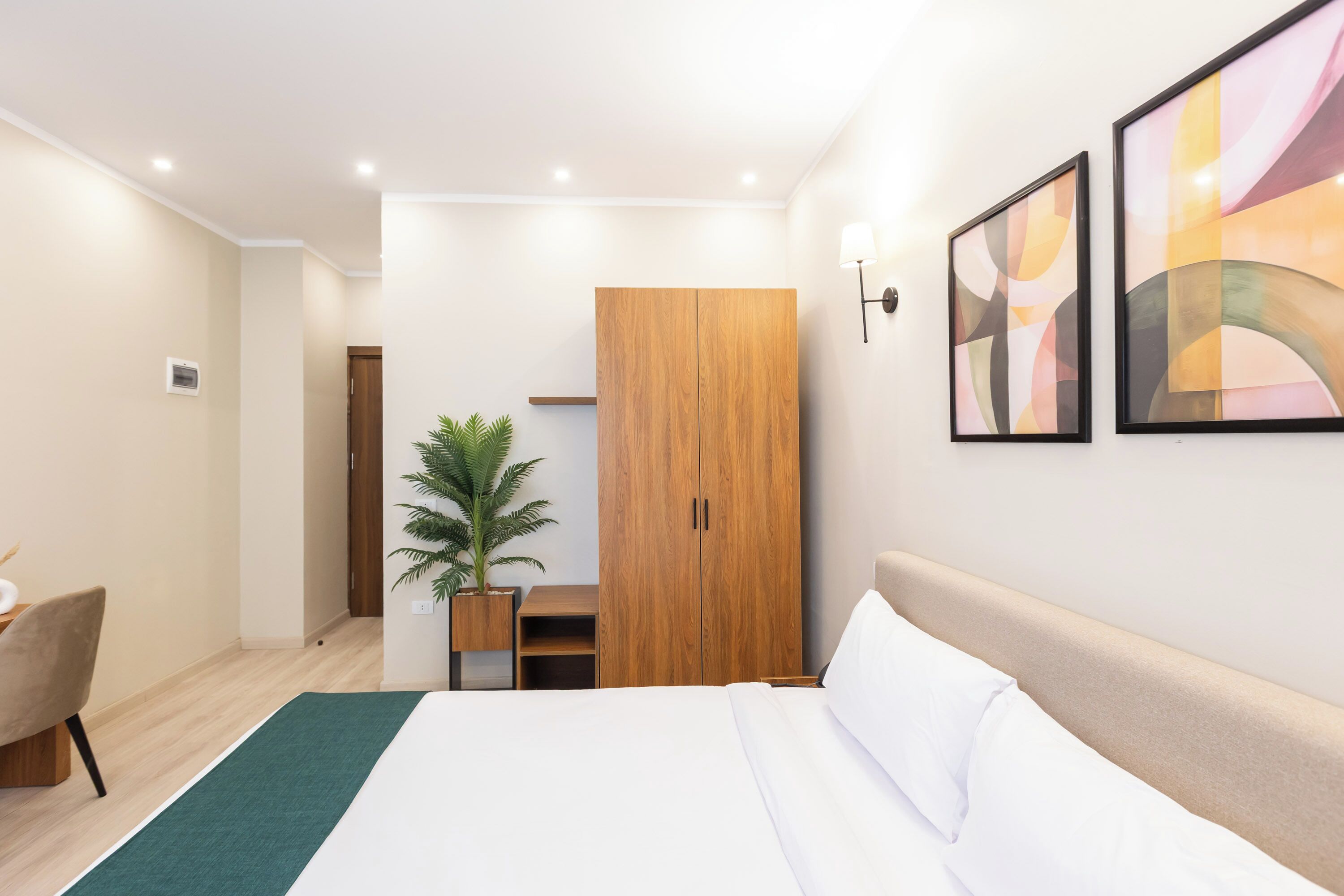 A modern hotel room with a large bed, a wooden wardrobe, a desk with a chair, and a potted plant.