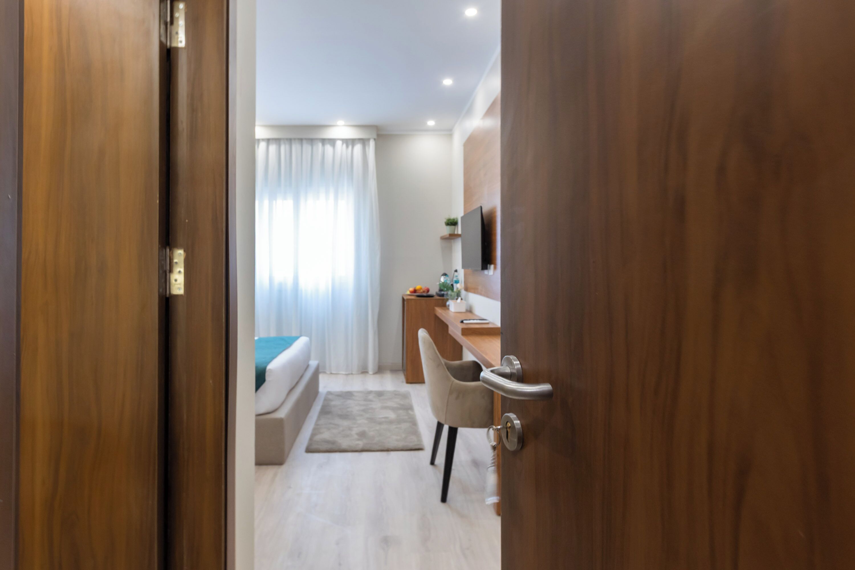 A modern hotel room with a wooden door, a bed, a desk, a chair, and a television on the wall.