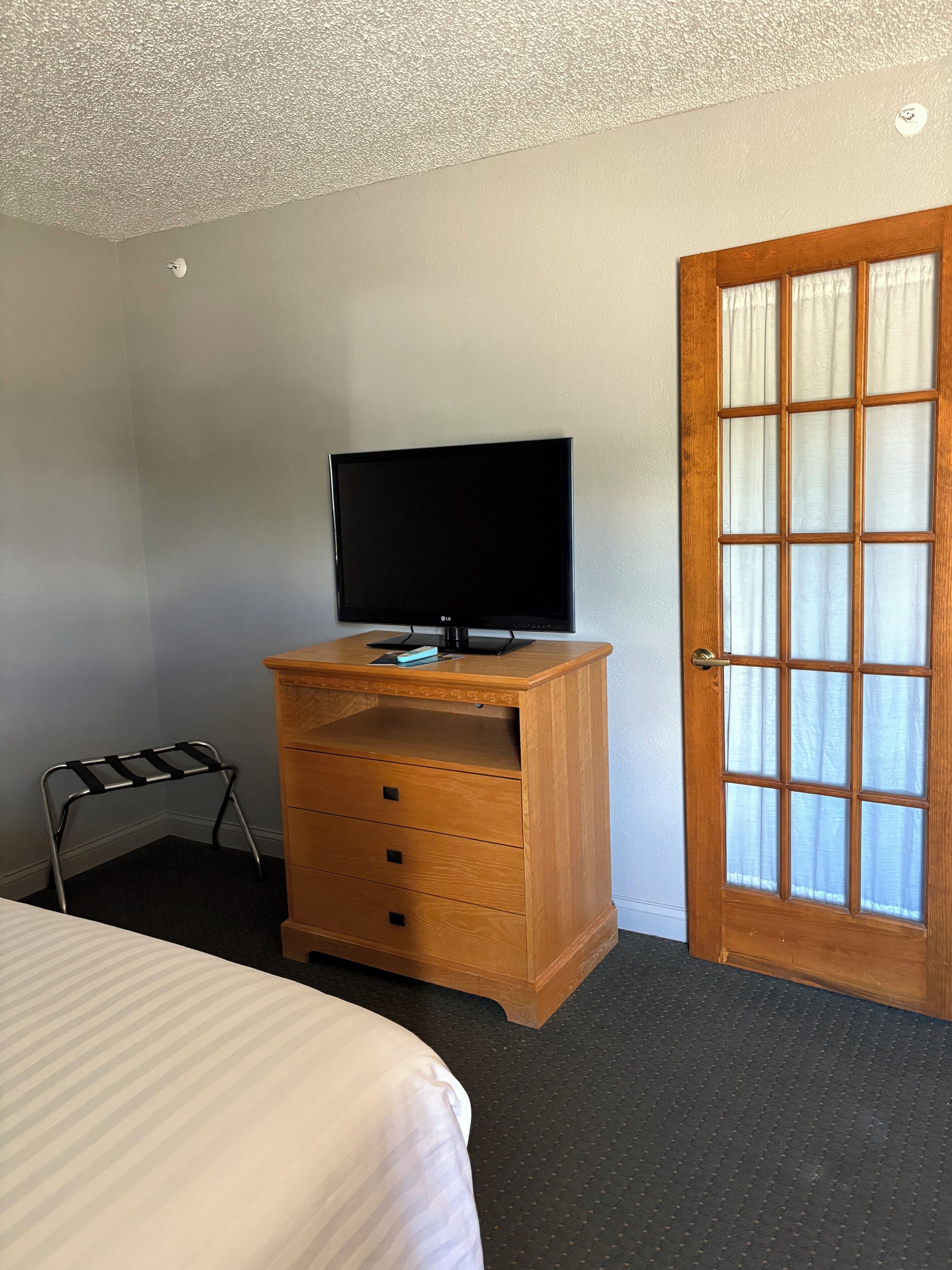Ein Hotelzimmer mit einem Bett, einer Holzkommode mit Fernseher, einem kleinen Metallgestell und einer Tür mit satinierten Glasscheiben.
