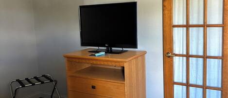 Ein Hotelzimmer mit einem Bett, einer Holzkommode mit Fernseher, einem kleinen Metallgestell und einer Tür mit satinierten Glasscheiben.