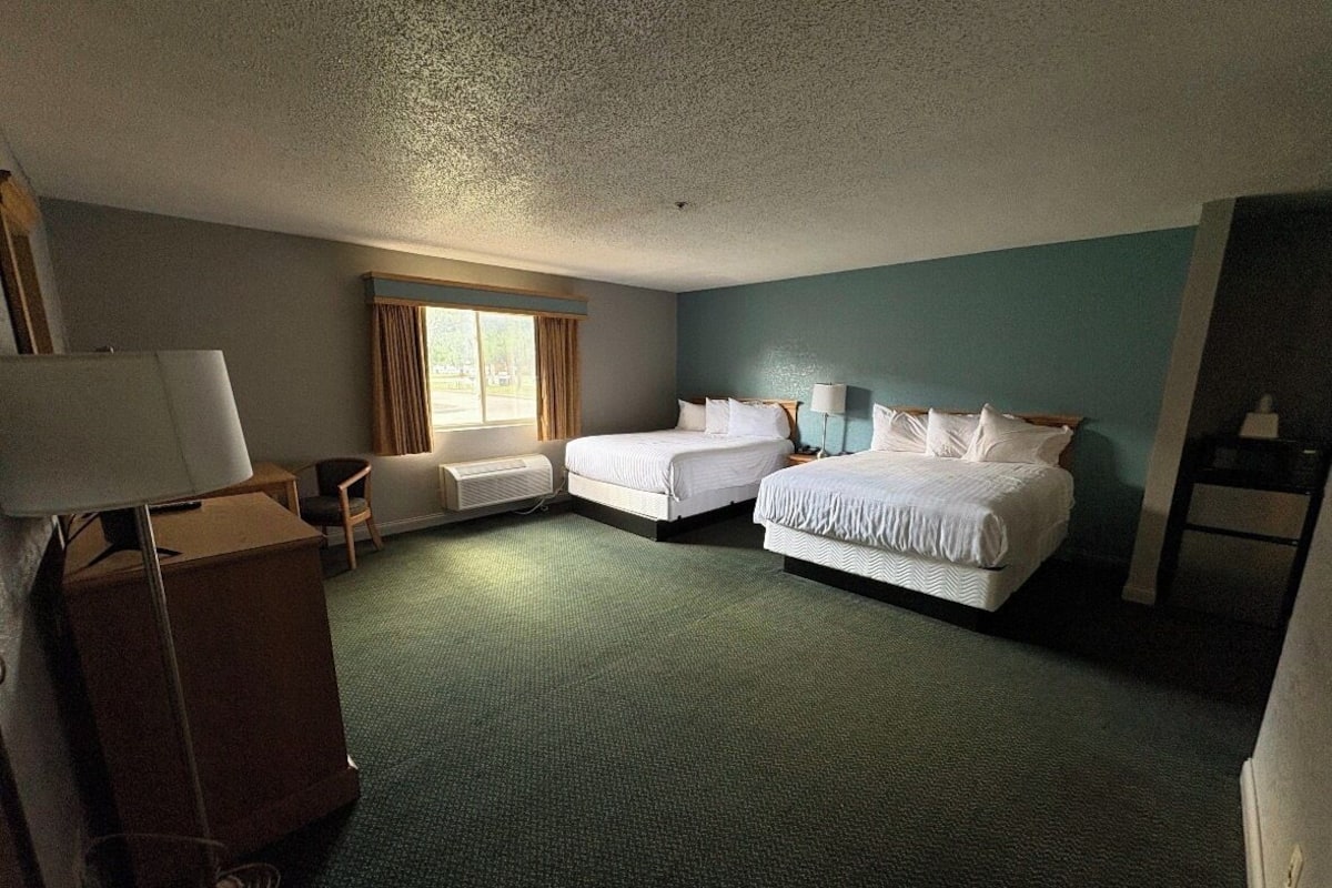 A hotel room with two beds, a desk, a chair, a lamp, and a window.
