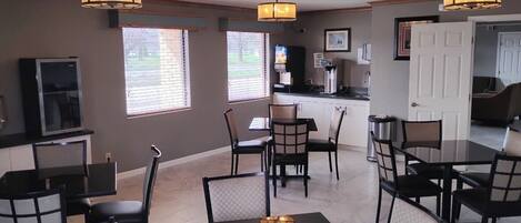 A dining area with tables and chairs, a kitchen in the background, and a door leading to another room.