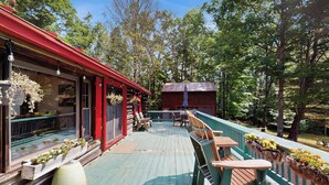 Een houten terras met buitenmeubilair, een rood gebouw en uitzicht op een bos.