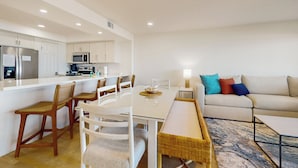 Una cocina moderna con armarios blancos, un refrigerador y un microondas. Hay un comedor con una mesa y sillas blancas, además de un sofá con almohadones de colores.