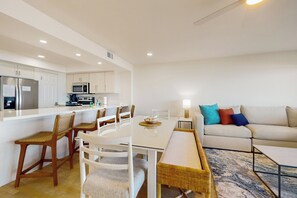 A modern kitchen with white cabinets, a refrigerator, and a microwave. There is a dining area with a white table and chairs, and a sofa with colorful pillows. - Sweeping View, Luxe Perks: Suntide III - Unit 708 (South Padre Island)