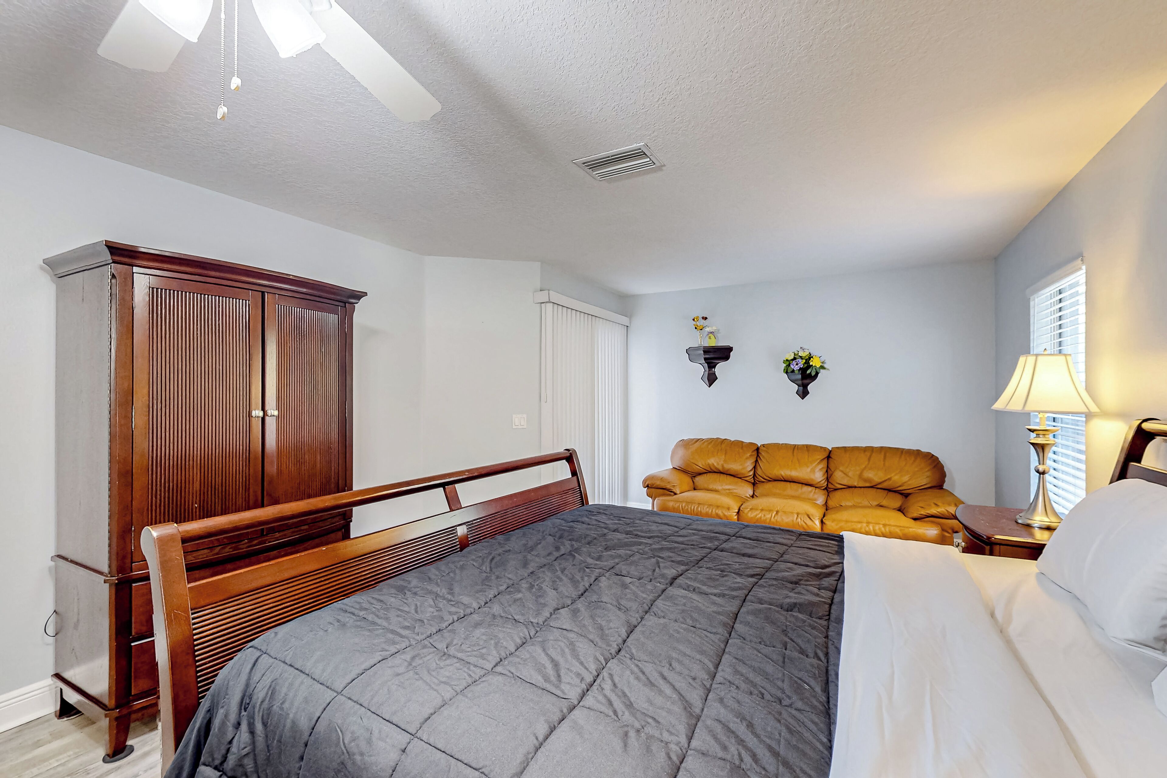 A bedroom with a bed, a wooden wardrobe, a leather sofa, a bedside table with a lamp, and a wall-mounted shelf with flowers.