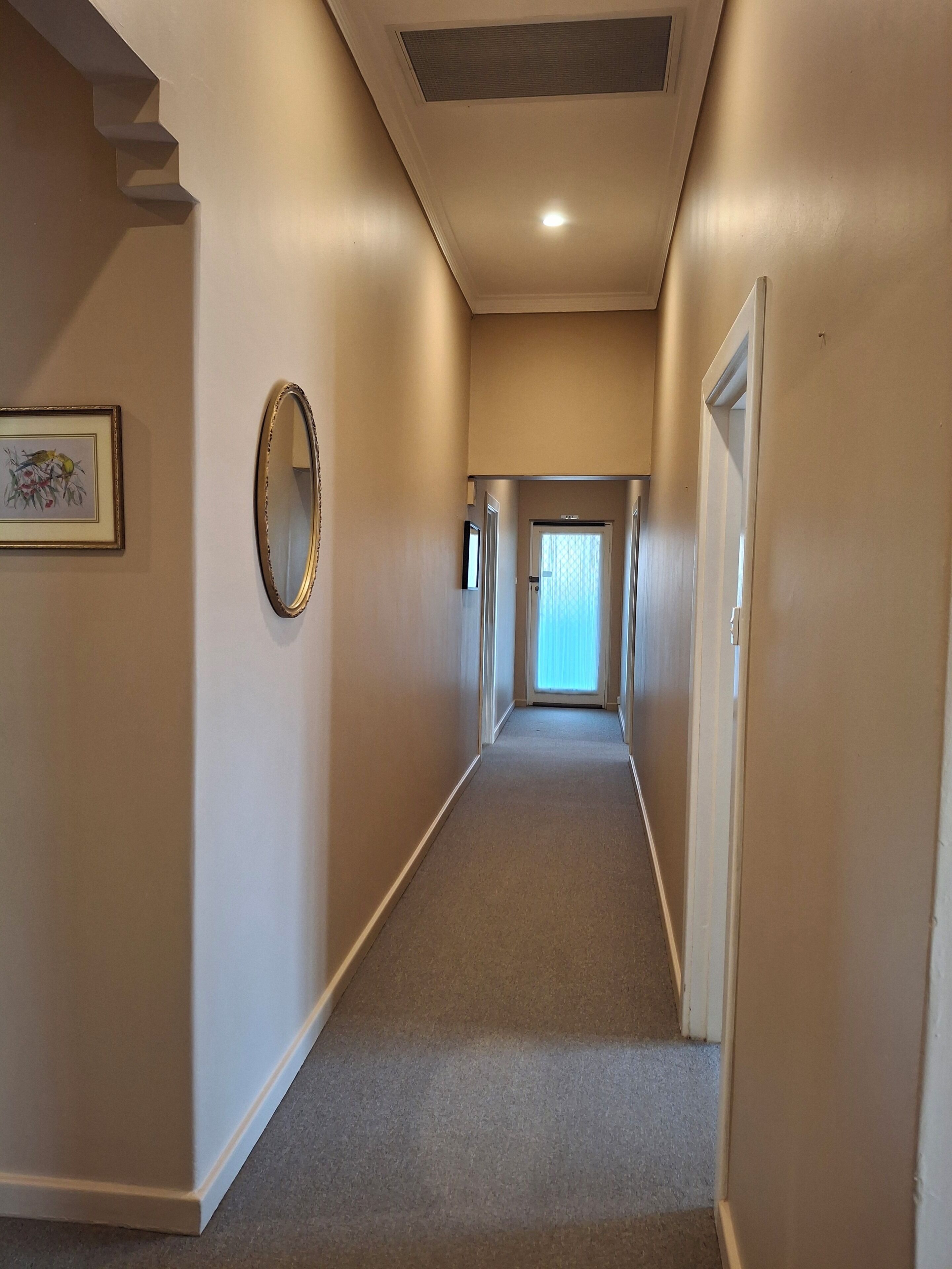 A long, narrow hallway with a carpeted floor, white walls, and a ceiling with recessed lighting. There are doors on both sides and a framed picture on the left wall.