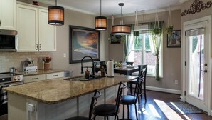 Une cuisine dotée d’un comptoir en granit, de tabourets de bar, de luminaires suspendus et d’une grande fenêtre agrémentée de rideaux.