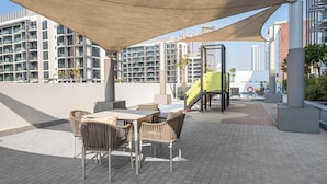 An outdoor patio area with a table and chairs, a children's play structure, and a large building in the background. - Stayshort - Studio Apartment at Azizi Riviera With Balcony (Dubai)